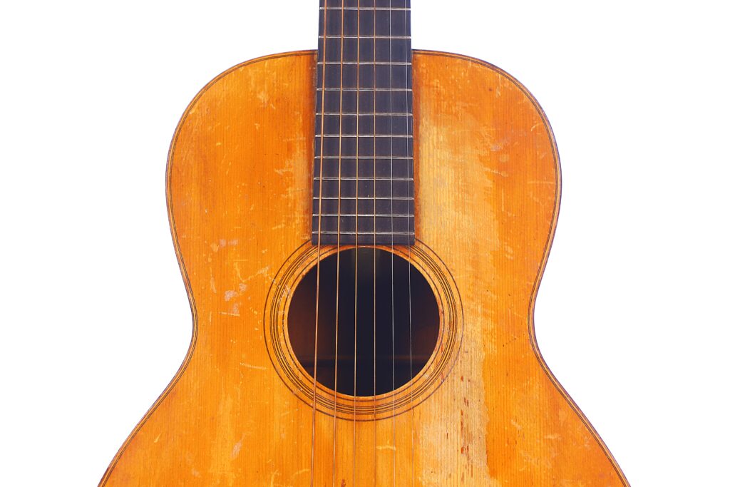 Close-Up Of A Martin 0-18 1926 Acoustic Guitar’s Body And Neck, Highlighting Its Well-Worn, Scratched Wooden Surface And Six Strings Against A White Background.