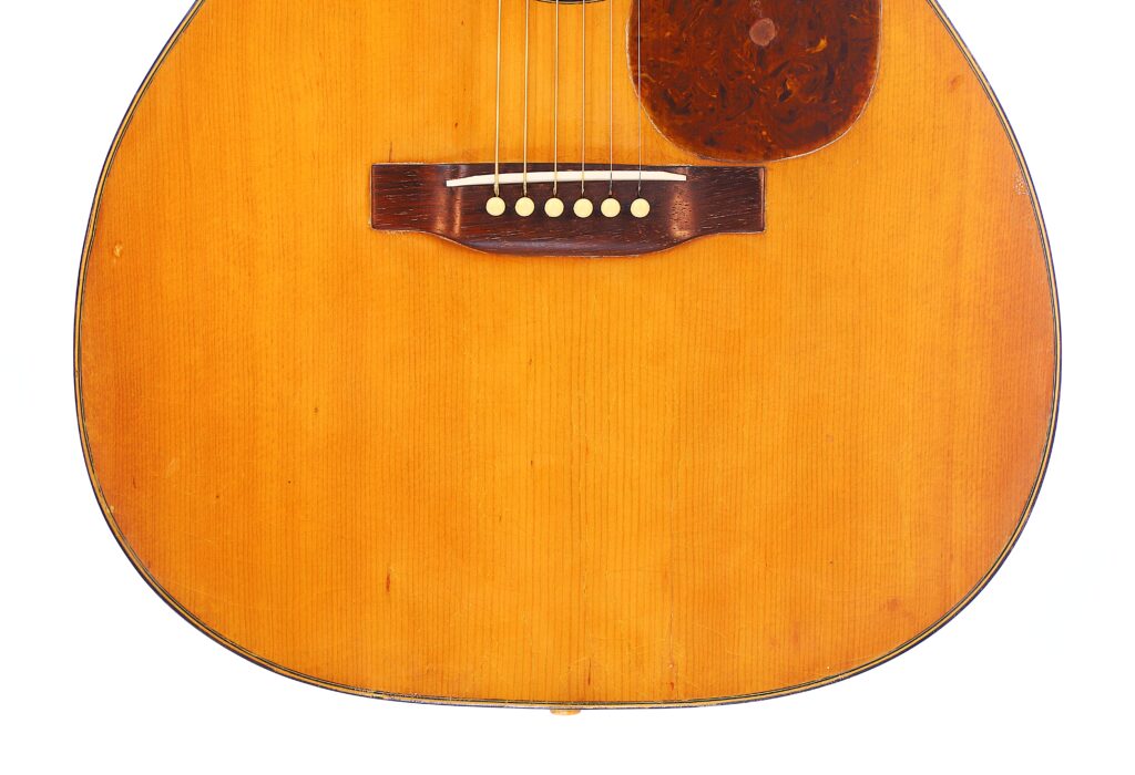 Close-Up Of The Martin 000-21 1947 Acoustic Guitar Body, Featuring Its Wooden Bridge, Six Strings, And Part Of The Pickguard Against A Natural Wood Grain Finish.