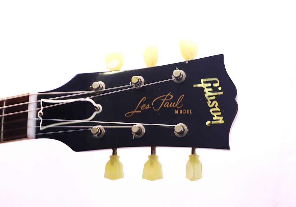 Close-Up Of The Headstock Of A Black Gibson Les Paul 1960 60Th Vos Anniversary Electric Guitar, Showing Tuning Pegs, Strings, And Gold Les Paul Model And Gibson Logos Against A White Background.