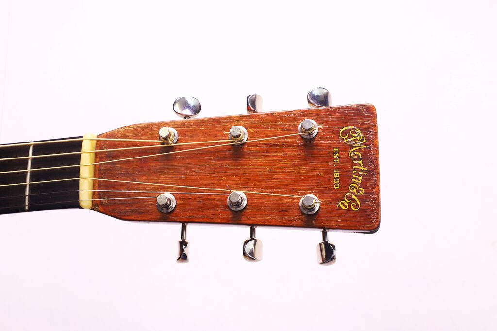 Close-Up Of The Headstock Of A Martin 000-21 1947 Acoustic Guitar, Displaying Six Tuning Pegs, Guitar Strings, And The Martin Logo On The Wood Surface Against A Plain White Background.