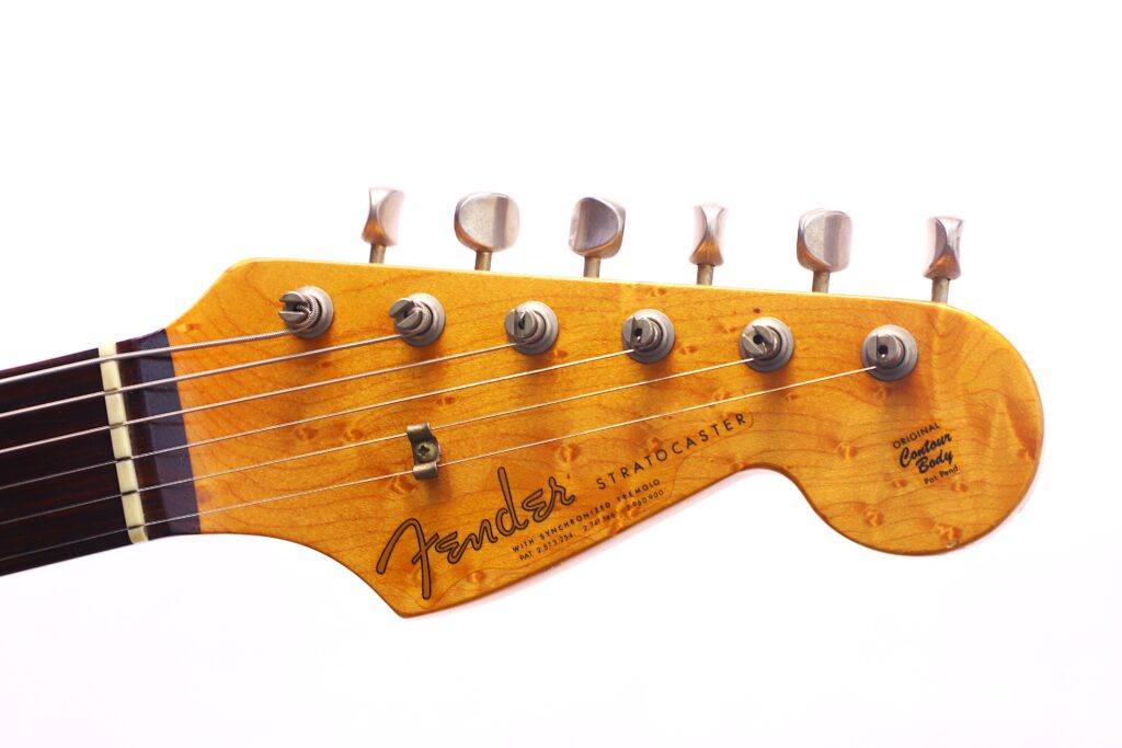Nahaufnahme Der Kopfplatte Einer Fender Stratocaster 1960 Relic Cunetto/Cruz E-Gitarre, Die Sechs Stimmwirbel, Saiten Und Das Fender-Logo Auf Holz Vor Einem Weißen Hintergrund Zeigt.