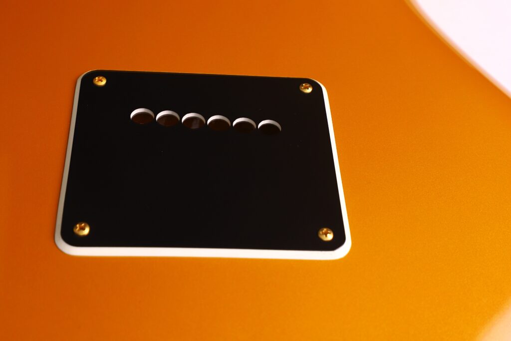 Close-Up Of The Back Of A Burns Legend 1994 Electric Guitar, Featuring An Orange Finish And A Black Rectangular Cover Plate With Six Holes Secured By Four Gold Screws.