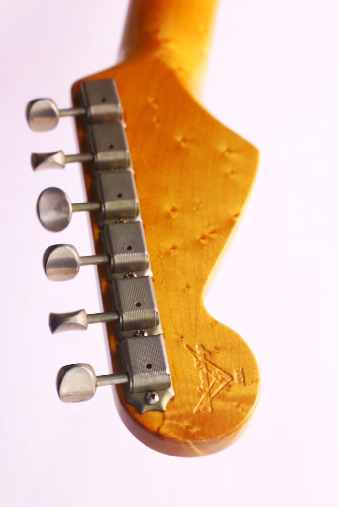 Nahaufnahme Der Kopfplatte Einer Fender Stratocaster 1960 Relic Cunetto/Cruz E-Gitarre Mit Sechs Silbernen Stimmwirbeln, Glänzender Holzoberfläche Und Einem Sichtbaren Gestempelten Logo Nahe Der Abgerundeten Kante Vor Einem Hellen Hintergrund.