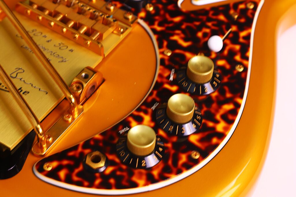 Close-Up Of A Burns Legend 1994 Electric Guitar Featuring An Orange And Black Tortoiseshell Pickguard, Gold Hardware, Three Black Control Knobs With White Numbers, And A White Switch Near The Knobs.