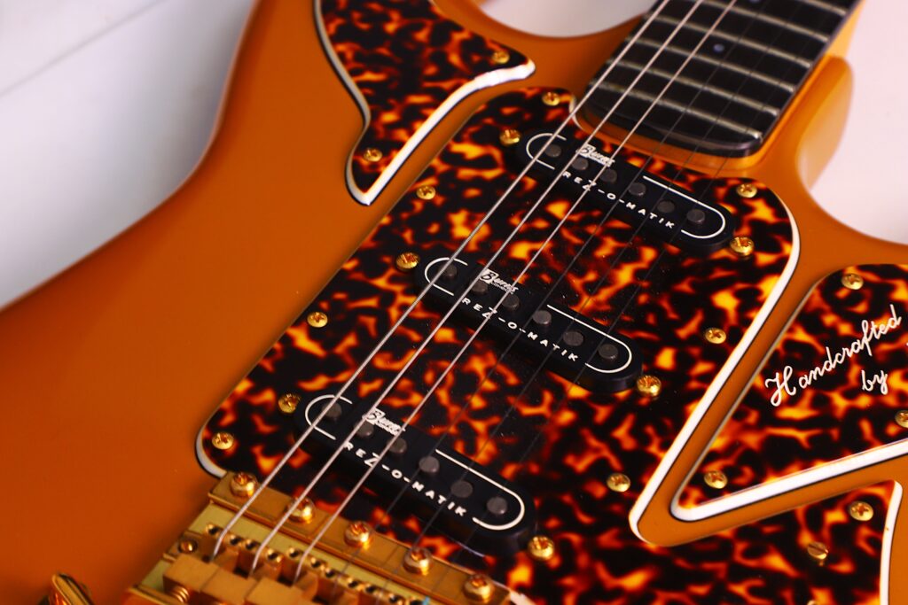 Close-Up Of A Burns Legend 1994 Electric Guitar Featuring An Orange Body, Gold Bridge, Tortoiseshell Pickguards, Three Single-Coil Pickups, And Part Of The Neck With Strings Visible.