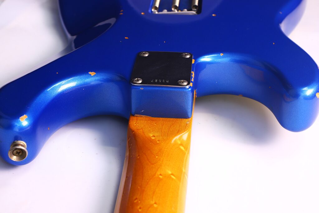 Nahaufnahme Der Rückseite Einer Fender Stratocaster 1960 Relic Cunetto/Cruz E-Gitarre Mit Einem Blauen, Farbabgesplitterten Korpus, Holzhals Und Einer Metallhalsplatte Mit Schrauben, Die Den Hals Mit Dem Korpus Verbinden.