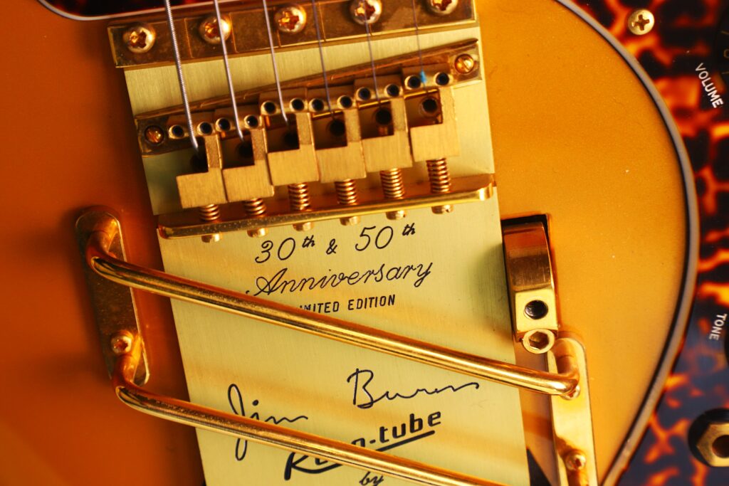 Close-Up Of A Gold Bridge And Tremolo System On The Burns Legend 1994 Electric Guitar, Featuring Engraved “30Th &Amp; 50Th Anniversary Limited Edition” Text And A Signature Plate, With Volume And Tone Knobs In The Background.