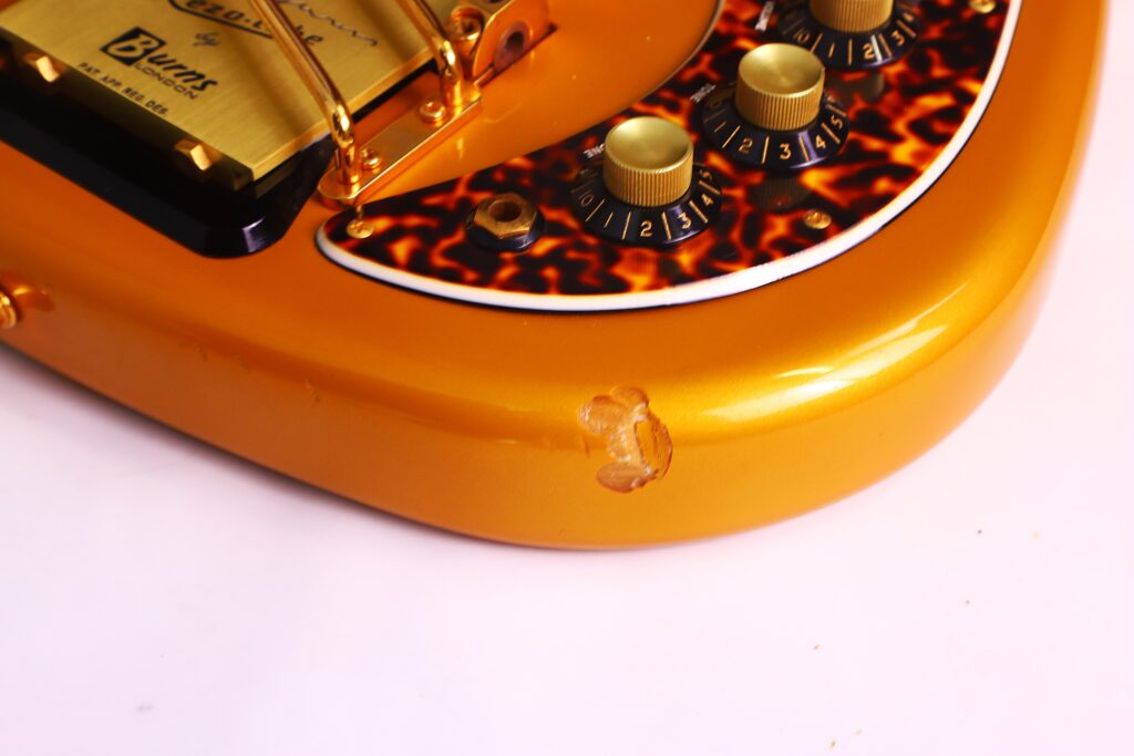 Close-Up Of A Burns Legend 1994 Electric Guitar In Gold, Featuring A Tortoiseshell Pickguard, Numbered Control Knobs, And A Visible Chip In The Paint On The Lower Edge Of The Body.