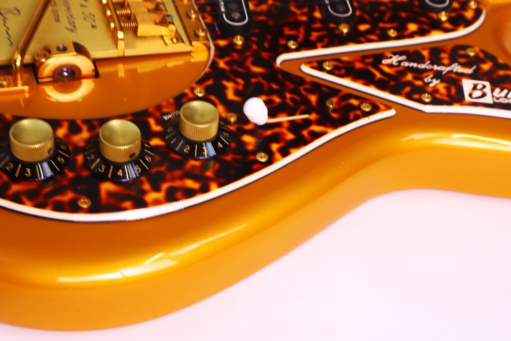 Close-Up Of A Burns Legend 1994 Electric Guitar Featuring A Golden-Orange Finish, Tortoiseshell Pickguard With &Quot;Handcrafted By&Quot; Partially Visible, Gold Hardware, Three Control Knobs, And A White Switch.