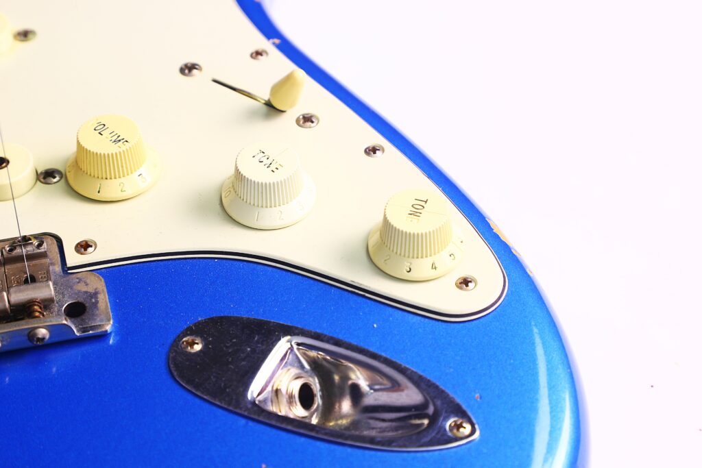 Nahaufnahme Der Regler Und Der Eingangsbuche An Einer Blauen Fender Stratocaster 1960 Relic Cunetto/Cruz E-Gitarre Mit Weißem Schlagbrett, Die Lautstärke- Und Klangregler Sowie Einen Teil Des Tonabnehmerwahlschalters Hervorhebt.