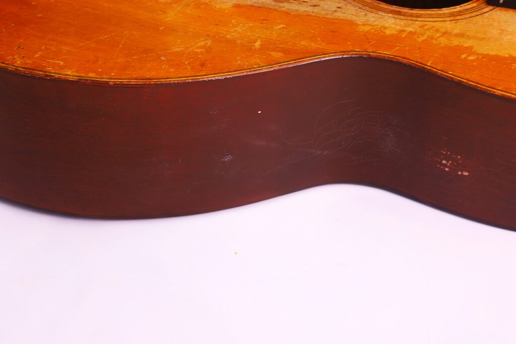 Close-Up Of The Side Of A Martin 0-18 1926 Acoustic Guitar, Highlighting Its Scratches And Age Marks, Set Against A Plain White Background.