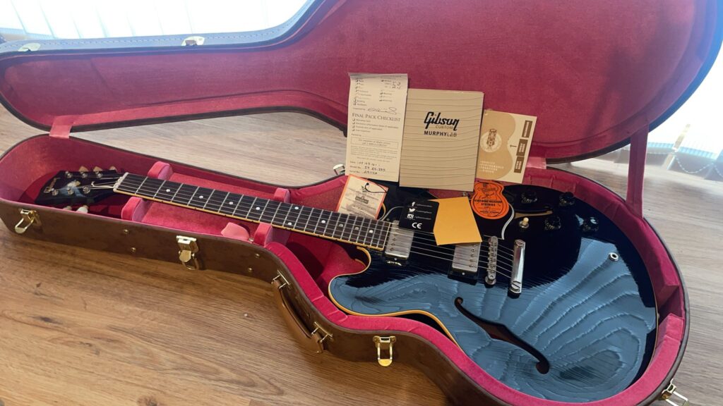 Eine Schwarze Gibson Es-335 Td Murphy Lab E-Gitarre, In Einem Braunen Hartschalenkoffer Mit Pinkfarbener Innenausstattung, Wird Auf Einer Holzoberfläche Präsentiert. Dokumente Und Etiketten Liegen Im Koffer Neben Der Gitarre.
