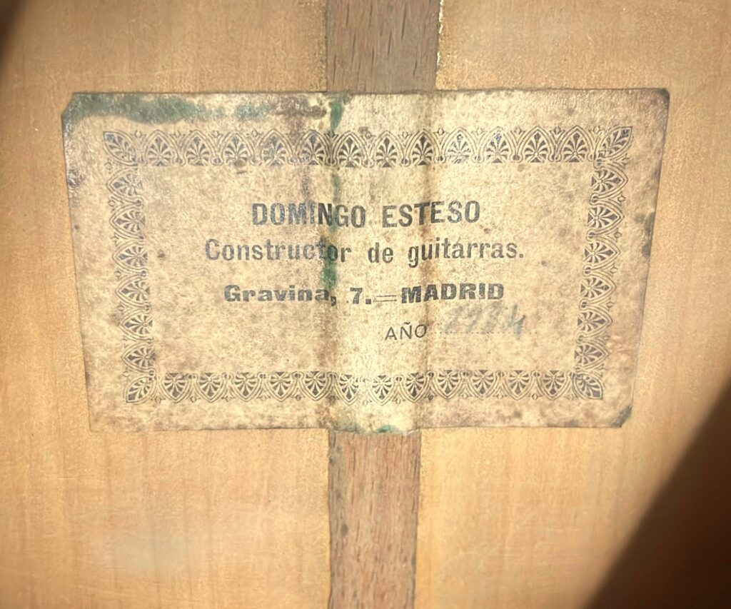 Uma fotografia de grande plano mostra uma etiqueta antiga no interior de uma guitarra clássica Domingo Esteso 1934 com a inscrição „Domingo Esteso, Constructor De Guitarras, Gravina, 7th - Madrid“, com o ano escrito à mão mas indistintamente anotado no bordo inferior.