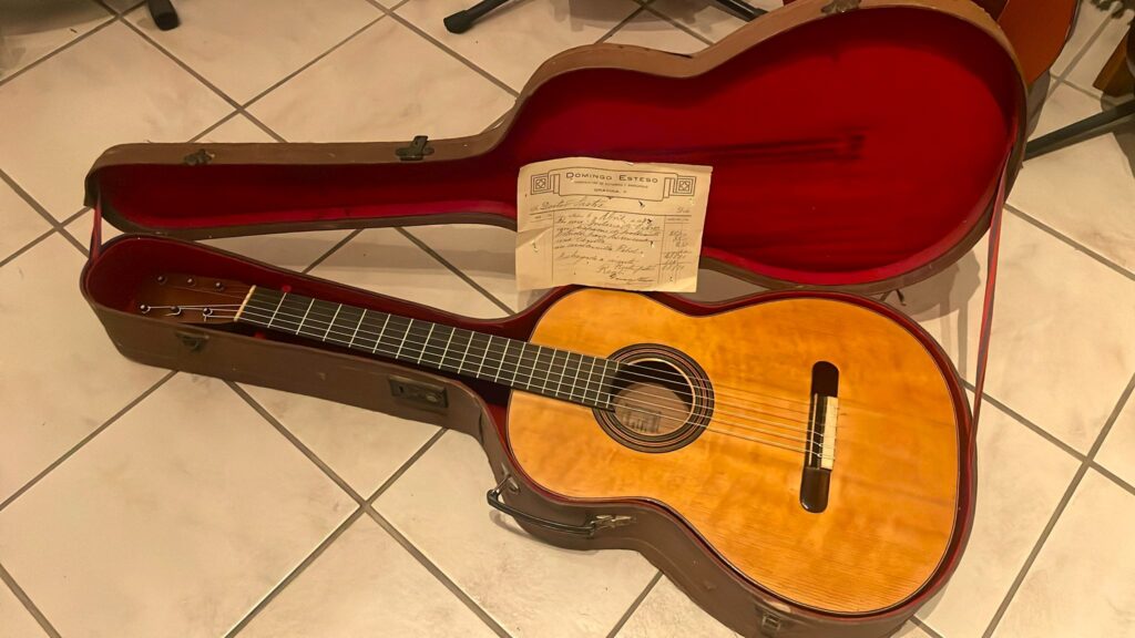 A guitarra clássica Domingo Esteso 1934 com acabamento em madeira clara é apresentada num estojo rígido aberto forrado com tecido vermelho. Um certificado encontra-se no topo, tudo colocado numa base de azulejos.