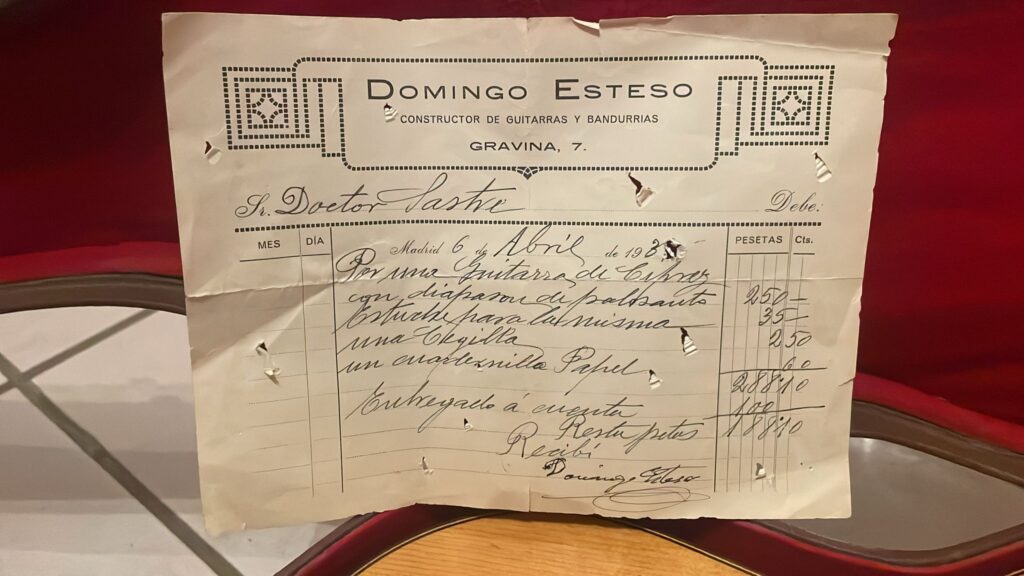 Um antigo recibo espanhol manuscrito, datado de 6 de abril de 1932, com uma lista de artigos em pesetas do fabricante de guitarras Domingo Esteso, encontra-se sobre uma almofada vermelha ao lado de uma guitarra clássica Domingo Esteso 1934.