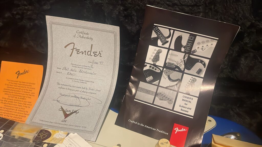 Ein Echtheitszertifikat Für Eine Fender Stratocaster 1960 Relic Cunetto/Cruz E-Gitarre, Ein Illustriertes Benutzerhandbuch, Weitere Dokumente Und Ein Teil Der Blauen Gitarre Werden Auf Einer Oberfläche Präsentiert.