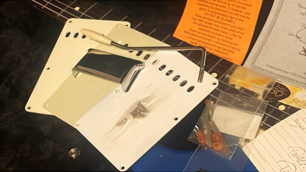 Verschiedene Gitarrenteile – Abdeckplatten, Schrauben, Ein Tremolohebel Und Kleinteile – Sind Zusammen Mit Dokumenten Und Verpackung Auf Einer Blauen Fender Stratocaster 1960 Relic Cunetto/Cruz E-Gitarre Ausgebreitet.