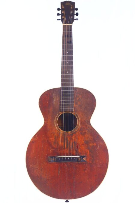 Die Gibson L-1 1926 Akustik Gitarre Hat Einen Abgenutzten Braunen Holzkorpus Mit Sichtbaren Kratzern, Ein Rundes Schallloch, Ein Dunkles Griffbrett Und Sechs Stimmwirbel, Die Vor Einem Weißen Hintergrund Abgebildet Sind. Die Gibson L-1 1926 Akustik Gitarre Hat Einen Abgenutzten Braunen Holzkorpus Mit Sichtbaren Kratzern, Ein Rundes Schallloch, Ein Dunkles Griffbrett Und Sechs Stimmwirbel, Die Vor Einem Weißen Hintergrund Abgebildet Sind.