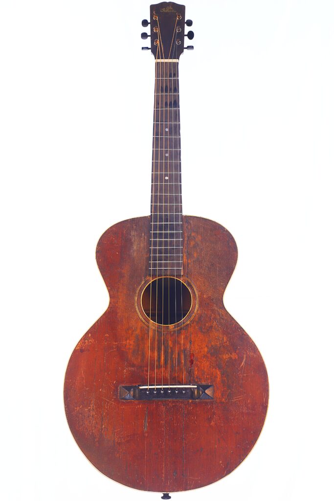 Die Gibson L-1 1926 Akustik Gitarre Hat Einen Abgenutzten Braunen Holzkorpus Mit Sichtbaren Kratzern, Ein Rundes Schallloch, Ein Dunkles Griffbrett Und Sechs Stimmwirbel, Die Vor Einem Weißen Hintergrund Abgebildet Sind.