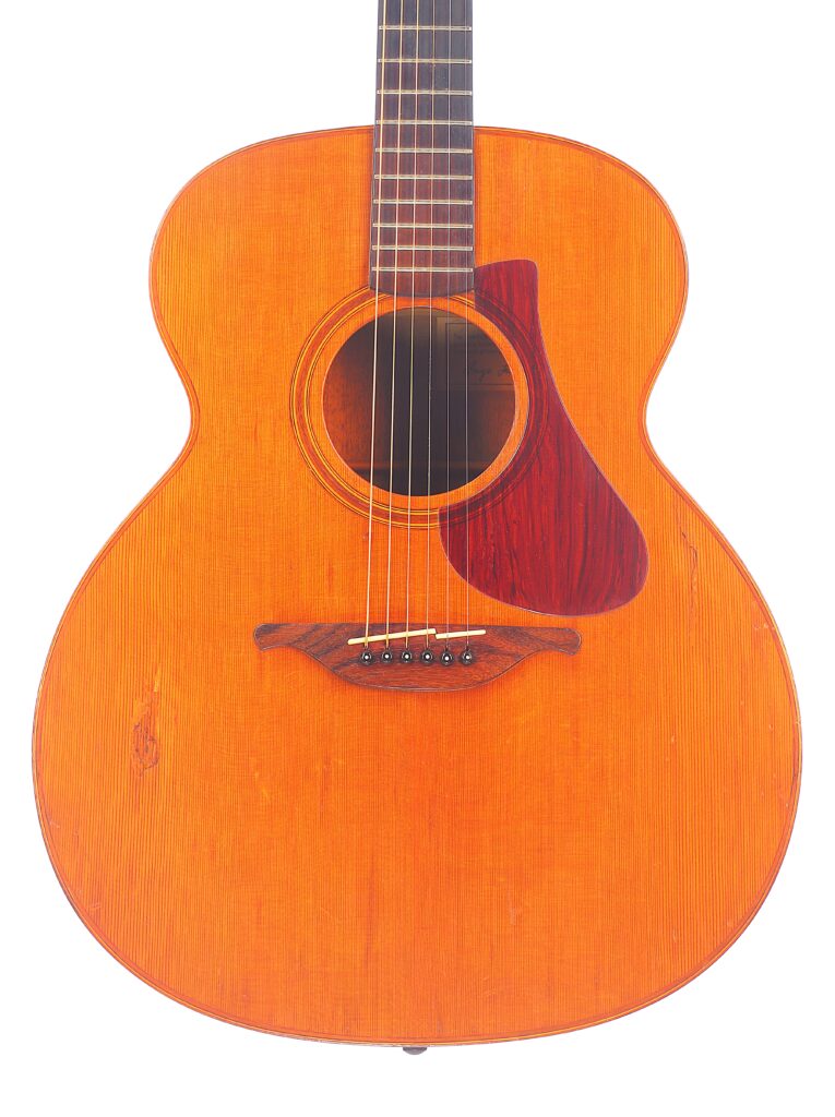 Die Lowden F-9 1980 Akustik Gitarre Zeichnet Sich Durch Ein Natürliches Holzfinish Mit Sichtbarer Maserung, Ein Rötlich-Braunes Schlagbrett Und Einen Markant Geformten Holzsteg Aus, Der Sich Vor Einem Weißen Hintergrund Präsentiert.