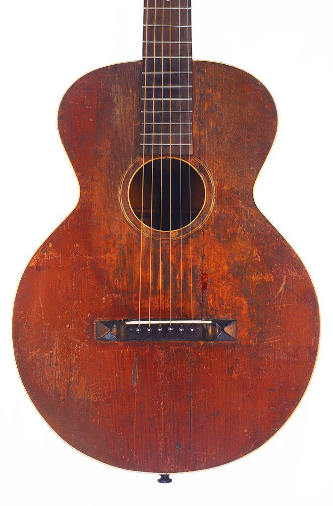 Eine Nahaufnahme Der Gibson L-1 1926 Akustik Gitarre Zeigt Den Zerkratzten, Rotbraunen Holzkorpus, Die Sechs Saiten Und Das Runde Schallloch, Fotografiert Vor Einem Schlichten Weißen Hintergrund.
