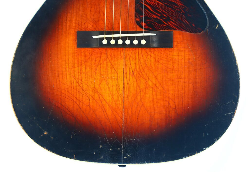 Nahaufnahme Des Korpus Der Gibson L-00 1934 Akustik Gitarre Mit Sunburst-Lackierung, Die Sechs Saiten, Einen Dunklen Steg Mit Weißen Stiften Und Sichtbare Oberflächenrisse Und Abnutzungen Zeigt.