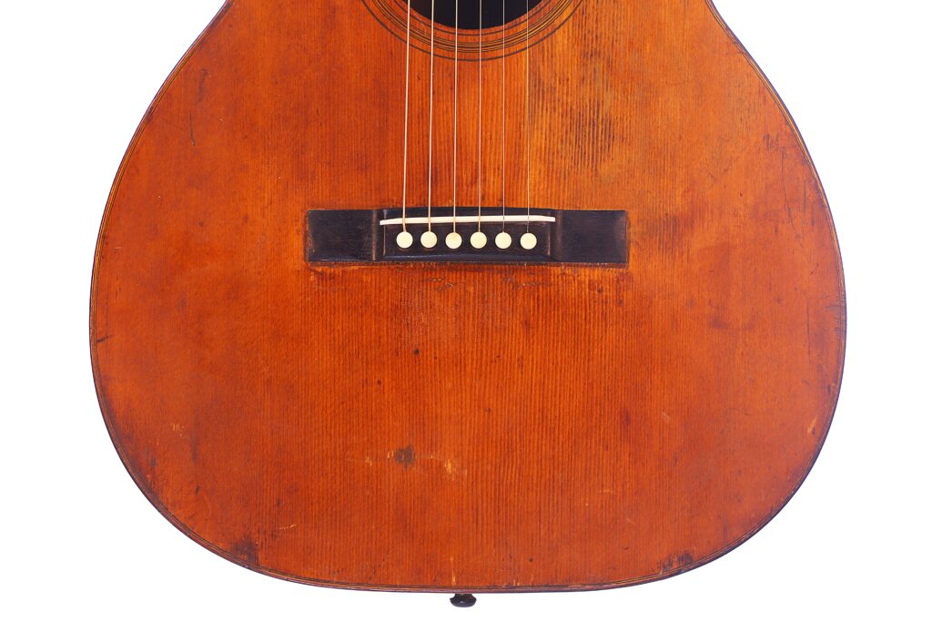 Close-Up Of A Martin 00-18 1922 Akustik Gitarre Body, Highlighting Its Warm Brown Wood Grain, Six Strings, Bridge, And Bridge Pins. Some Scratches Are Visible On The Wooden Surface.