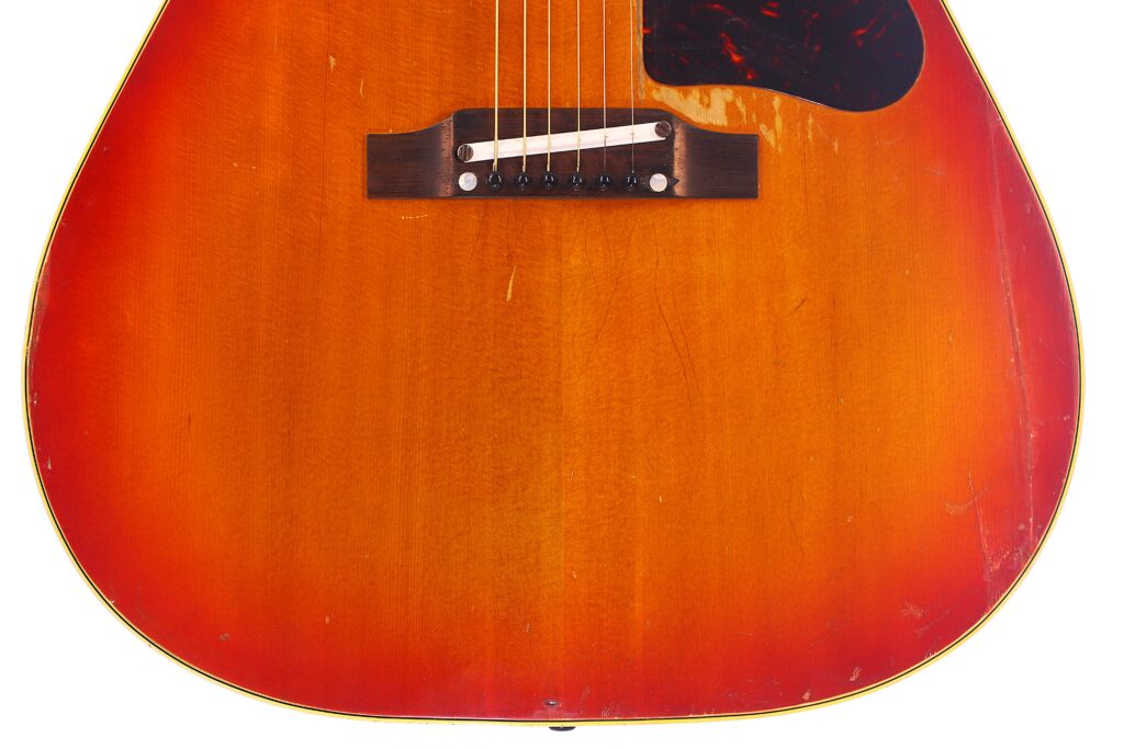 Nahaufnahme Des Korpus Einer Gibson J-45 1962 Akustik Gitarre Mit Sunburst-Finish, Die Den Steg, Die Saiten Und Das Schlagbrett Zeigt. Das Holz Weist Einen Warmen Orange-Roten Farbverlauf Mit Sichtbaren Kratzern Und Gebrauchsspuren Auf.