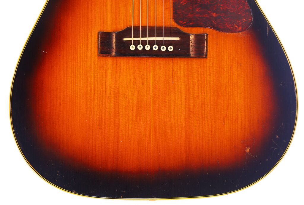 Nahaufnahme Der Unteren Hälfte Einer Gibson J-45 1957 Akustik-Gitarre Mit Sunburst-Lackierung, Die Den Steg, Die Sechs Saiten, Das Schlagbrett Und Einen Teil Des Korpus Vor Einem Weißen Hintergrund Zeigt.