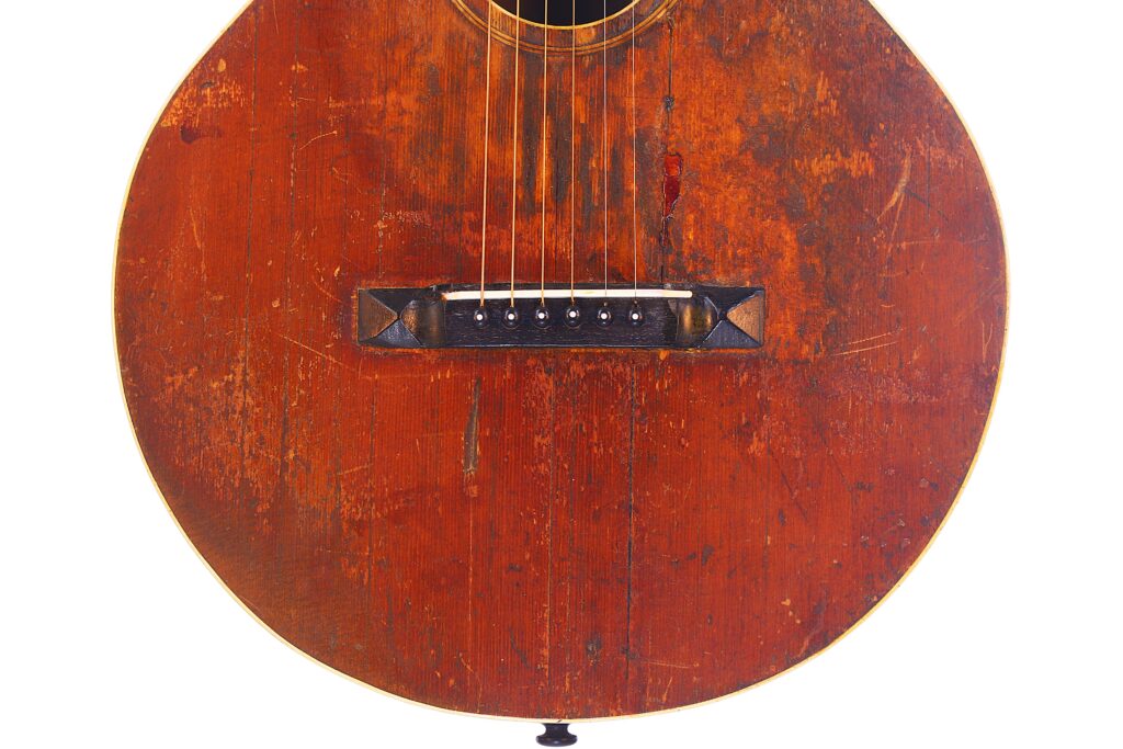 Eine Nahaufnahme Des Korpus Der Gibson L-1 1926 Akustik Gitarre Zeigt Sichtbare Kratzer, Schrammen Und Eine Dunkle Holzbrücke Mit Sechs Saiten.