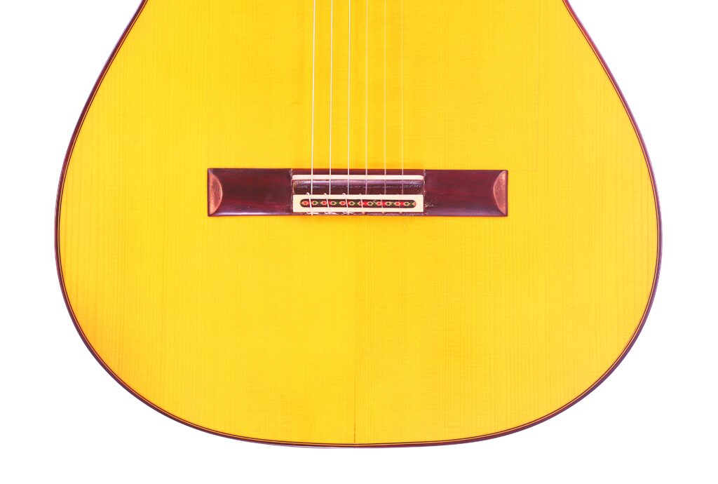 Nahaufnahme Des Gelben Korpus Der Graciliano Perez 2025 Flamenco-Gitarre Mit Sechs Saiten Und Einem Dunkelbraunen Holzsteg, Beschnitten, Um Nur Den Unteren Teil Vor Einem Weißen Hintergrund Zu Zeigen.