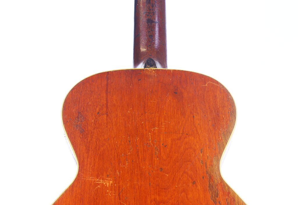 Eine Nahaufnahme Zeigt Die Rückseite Einer Gibson L-1 1926 Akustik-Gitarre Vor Einem Weißen Hintergrund, Wobei Die Abgenutzte Oberfläche, Sichtbare Kratzer Und Der Nach Oben Ragende Hals Hervorgehoben Werden.