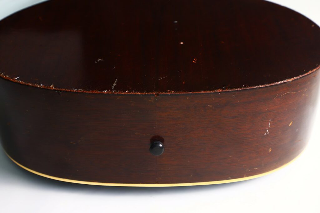 Nahaufnahme Der Seite Einer Gibson L-00 1934 Akustikgitarre Mit Dunklem Finish Mit Sichtbaren Kratzern Und Dellen, Cremefarbenem Binding Entlang Der Kante Und Einem Mittig Platzierten Gurtknopf.