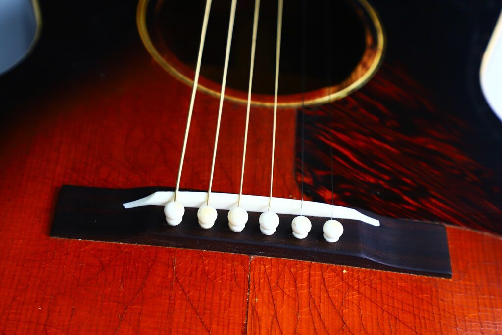 Nahaufnahme Des Stegs Einer Gibson L-00 Akustikgitarre Von 1934. Zu Sehen Sind Sechs An Weißen Stegstiften Befestigte Saiten Auf Einem Rötlich-Braunen Holzkörper Mit Sichtbarer Maserung Und Einem Teil Des Schalllochs.