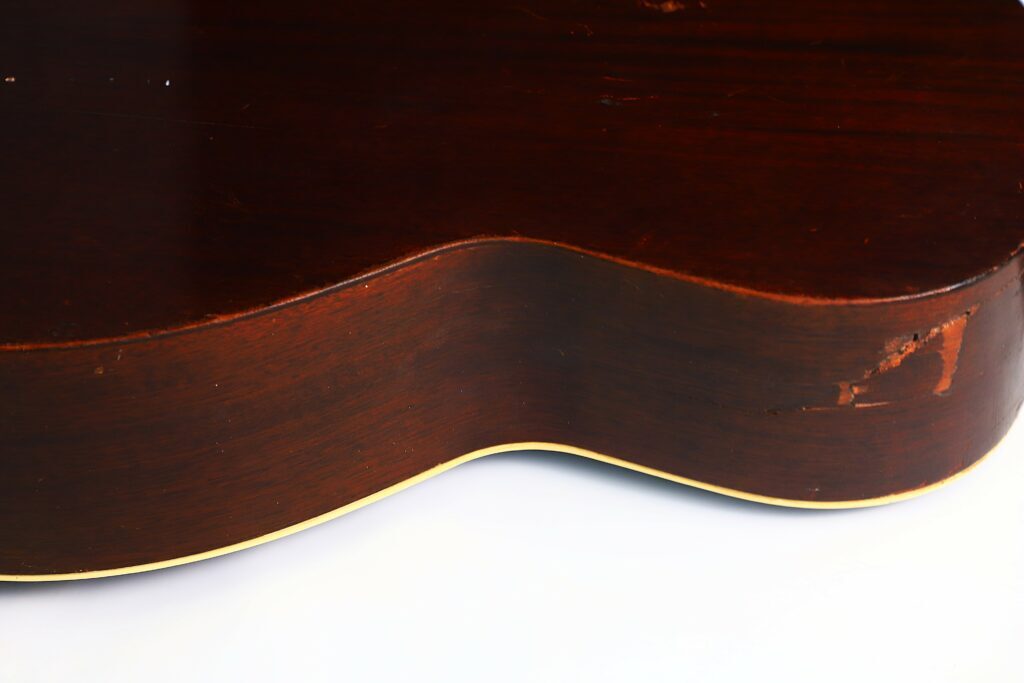 Nahaufnahme Der Seite Einer Gibson L-00 1934 Akustik-Gitarre Aus Dunkelbraunem Holz, Die Einen Sichtbaren Kratzer Und Eine Abgeplatzte Stelle Am Korpus Zeigt, Der Auf Einer Weißen Oberfläche Ruht.