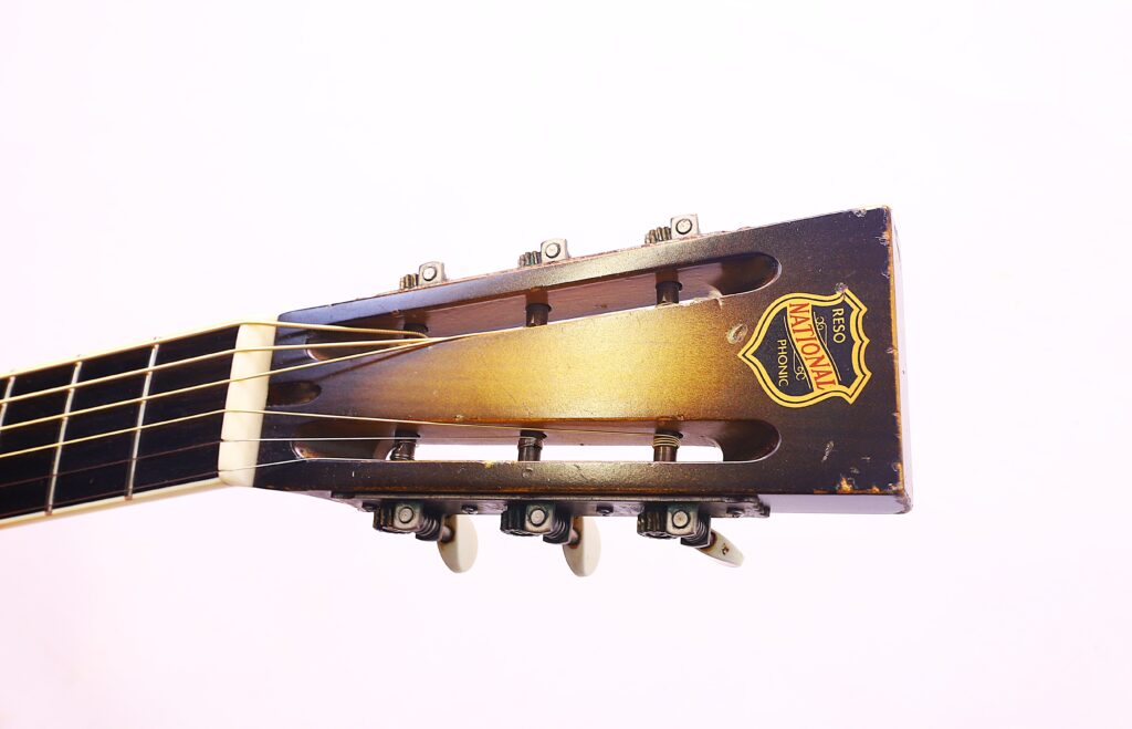 Nahaufnahme Der Kopfplatte Einer National Reso-Phonic Style O 2009 Akustikgitarre, Die Sechs Stimmwirbel Und Ein Gelbes Und Rotes National-Logo Vor Einem Schlichten Weißen Hintergrund Zeigt.