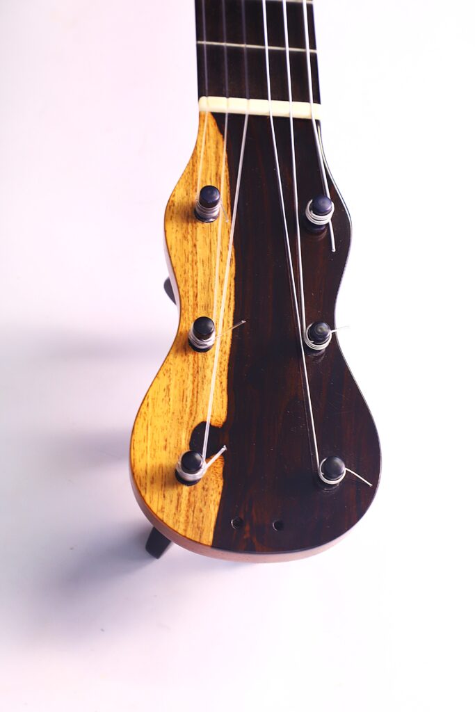 Nahaufnahme Der Kopfplatte Der Grobert Romantikgitarre 2016 Von Yoann Charbonnier, Gefertigt Aus Zwei Kontrastierenden Hölzern, Mit Vier Schwarzen Stimmwirbeln Und Metallsaiten, Vor Einem Schlichten Weißen Hintergrund.