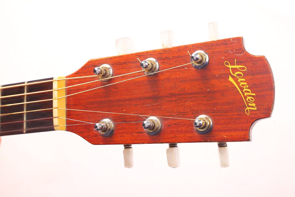 Eine Nahaufnahme Der Kopfplatte Einer Lowden F-9 1980 Akustikgitarre Zeigt Sechs Stimmwirbel, Sichtbare Saiten Und &Quot;Lowden&Quot; In Gelber Schrift Auf Holz Vor Einem Schlichten Weißen Hintergrund.