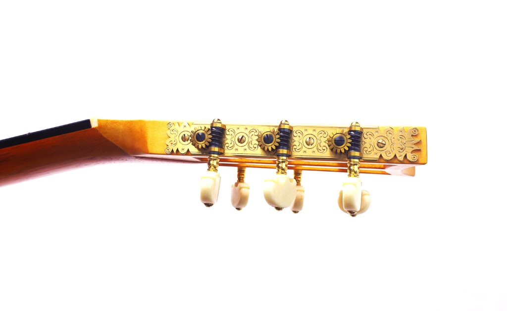 흰색 바탕에 세 개의 흰색 튜닝 페그와 장식 조각이 새겨진 화려한 황금색 튜너 플레이트가 있는 베른하르트 크레세 1999 파노르모 로맨틱 기타의 헤드스톡 클로즈업.