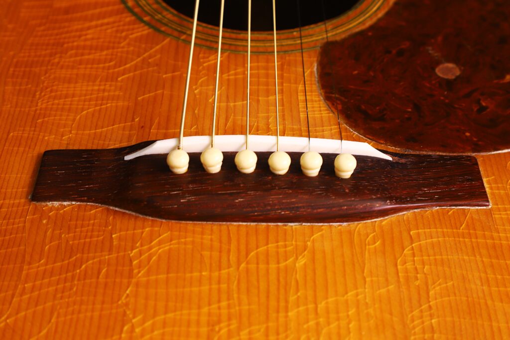 Nahaufnahme Des Stegs Und Der Sechs Saiten Einer Martin 000-21 Akustikgitarre Von 1947 Mit Weißen Stegstiften Im Holzsteg Und Dem Glänzenden, Strukturierten Korpus Der Gitarre.