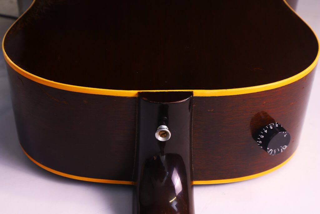 Nahaufnahme Der Rückseite Einer Gibson J-45 1957 Akustik Gitarre, Wo Der Hals Auf Den Korpus Trifft. Hervorzuheben Sind Das Dunkle Holz, Die Gelbe Zierleiste, Der Gurtknopf Und Der Schwarze Lautstärkeregler.