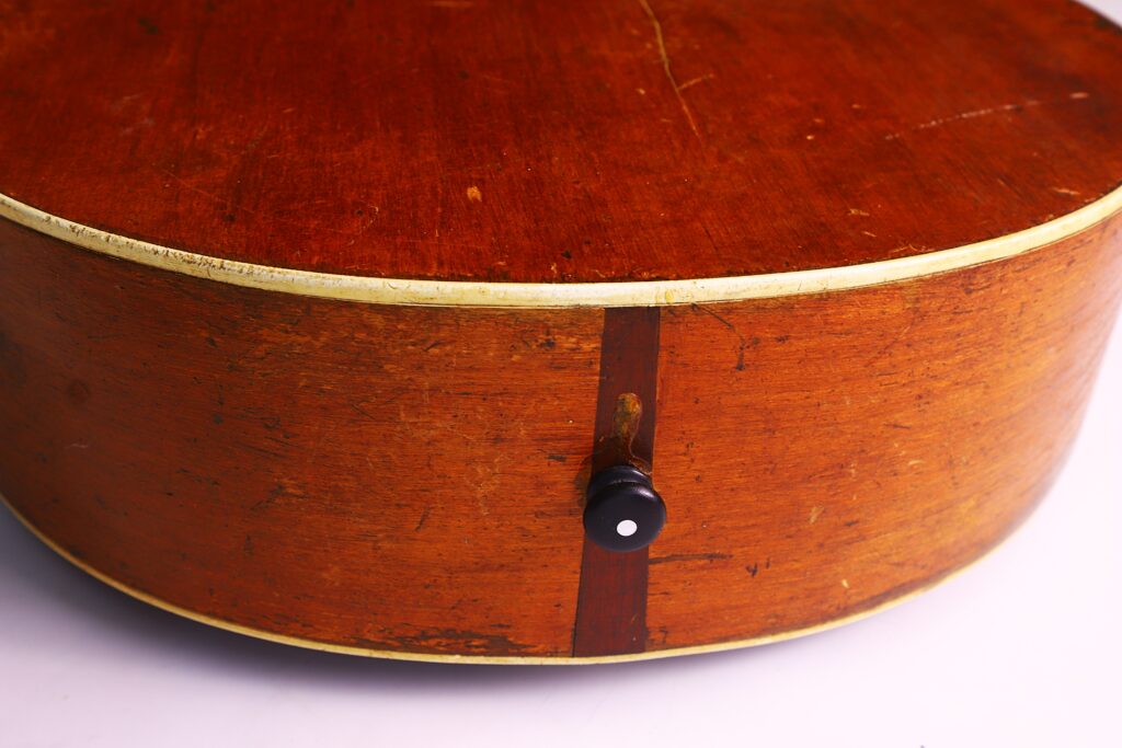 Nahaufnahme Der Gibson L-1 1926 Akustik-Gitarre, Die Das Abgenutzte Finish, Die Cremefarbene Randeinfassung, Den Schwarzen Gurtknopf Und Eine Sichtbare Vertikale Holznaht Hervorhebt.