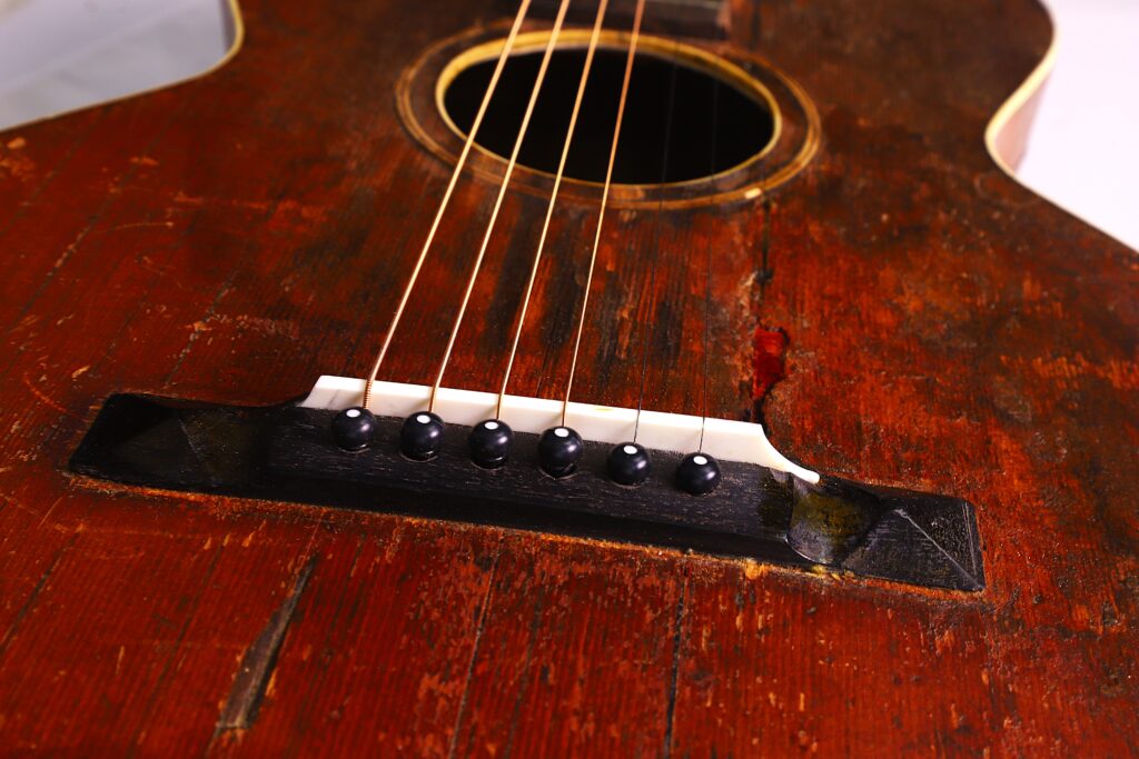 Nahaufnahme Einer Gibson L-1 1926 Akustik-Gitarre Mit Schwerpunkt Auf Dem Steg, Den Saiten Und Dem Schallloch. Die Zerkratzte Holzoberfläche Unterstreicht Den Vintage-Charme Der Gitarre.