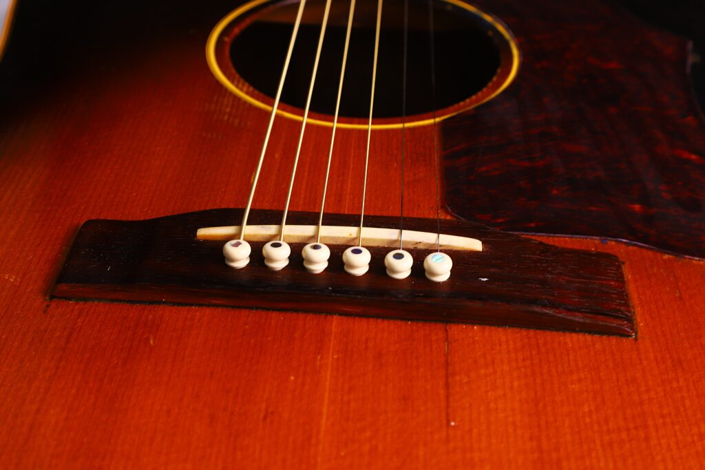 Nahaufnahme Des Stegs Der Gibson J-45 1957 Akustik Gitarre Mit Sechs Saiten, Weißen Stegstiften Und Einem Warmen Braunen Holzkörper. Das Schallloch Und Ein Teil Des Schlagbretts Sind Ebenfalls Zu Sehen.