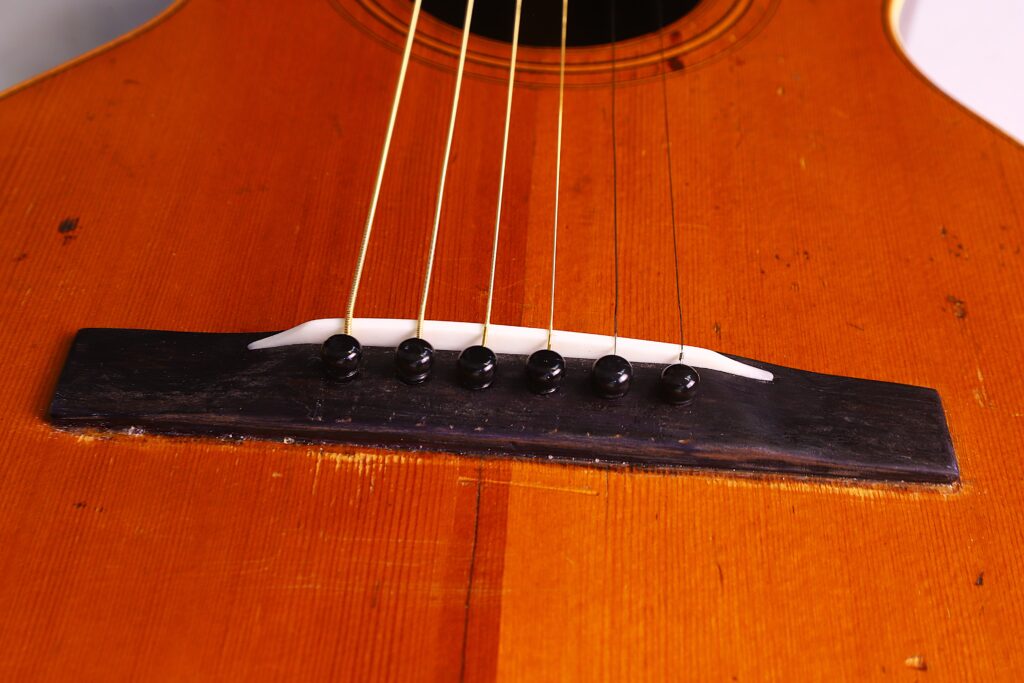 Nahaufnahme Des Stegs Der Washburn 1894 Parlor Akustik Gitarre Mit Sechs Saiten, Die Mit Schwarzen Stegstiften Auf Einem Warmen Braunen Holzkorpus Verankert Sind.