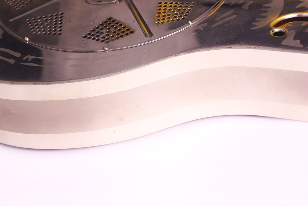 Nahaufnahme Einer National Reso-Phonic Style O 2009 Akustik Gitarre, Die Den Metallischen Korpus Mit Glänzender, Reflektierender Oberfläche Und Runder, Gelochter Deckplatte Vor Einem Hellen Hintergrund Zeigt. Nur Ein Teil Der Gitarre Ist Sichtbar.