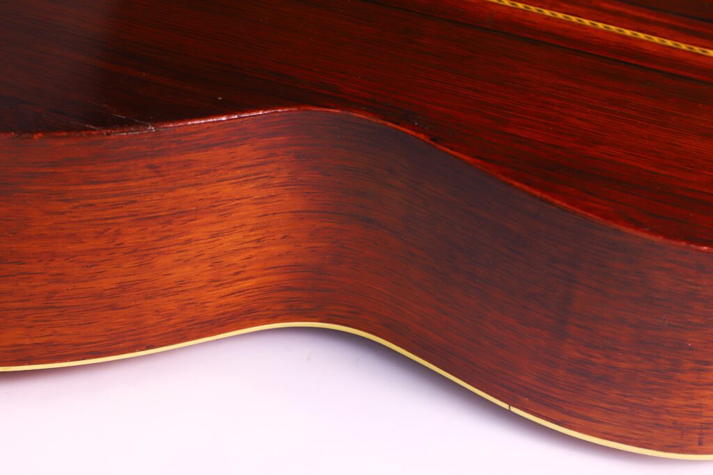 Nahaufnahme Der Geschwungenen Holzseite Der Washburn 1894 Parlor Akustik Gitarre. Hervorzuheben Sind Die Reiche Braune Maserung, Die Glänzende Oberfläche Und Die Dünne Cremefarbene Zierleiste Entlang Der Kante Vor Einem Weißen Hintergrund.