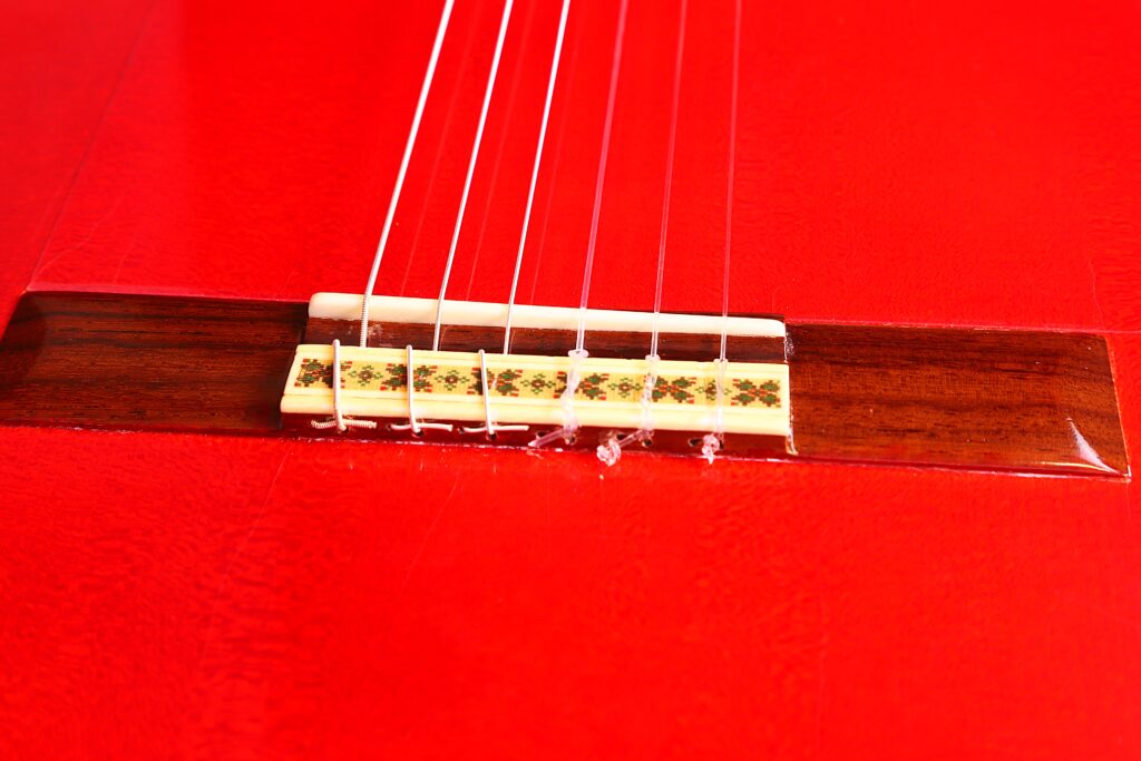Nahaufnahme Des Stegs Und Der Saiten Der Manuel Guillena 2017 Blanca Flamenco Gitarre Auf Einem Leuchtend Roten Korpus, Wobei Die Detaillierten Einlegearbeiten Am Steg Und Die Glänzende, Glatte Oberfläche Der Gitarre Hervorgehoben Werden.