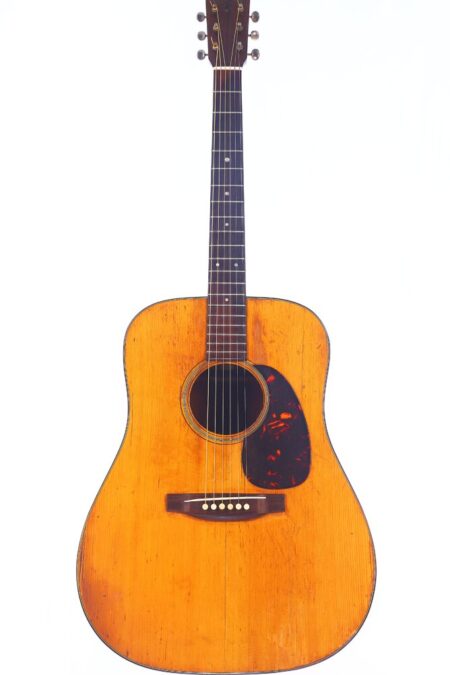 A Martin D-18 1965 Acoustic Guitar With A Natural Wood Finish, Dark Pickguard, And Six Strings Stands Upright Against A White Background. The Body Shows Visible Wear And Markings, Highlighting Its Vintage Appeal.