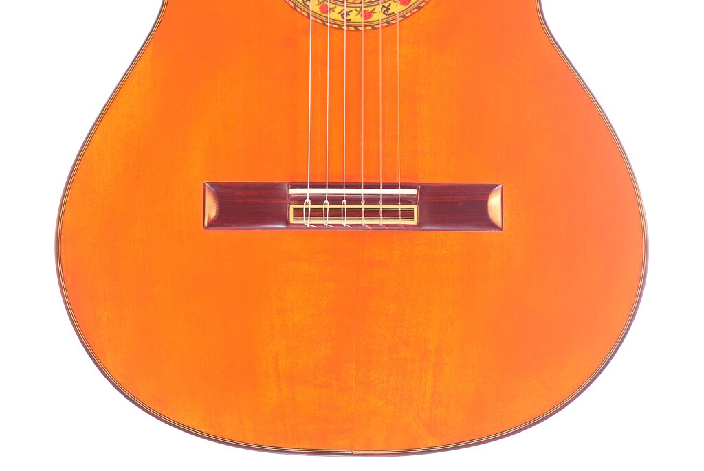 Nahaufnahme Des Korpus Der Pedro Fernández Luque 2020 Blanca Flamenco Gitarre Mit Sechs Saiten, Einem Holzsteg, Einer Glänzenden Orange-Braunen Lackierung Und Einem Teil Des Dekorativen Schalllochs An Der Decke.